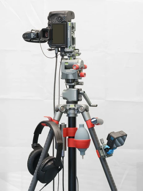 Professional video camera mounted on a red and gray tripod with monitor arm and wireless receiver attached.