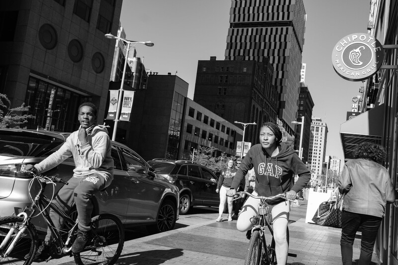 Two cyclists riding bikes on an urban street with parked cars and tall buildings in the background.