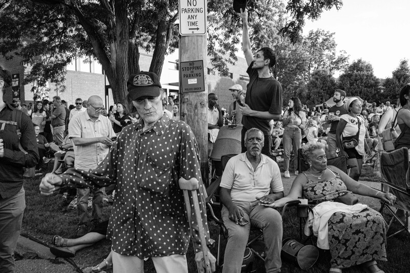 Black and white photograph of a crowded outdoor gathering with people seated and standing beneath large trees.