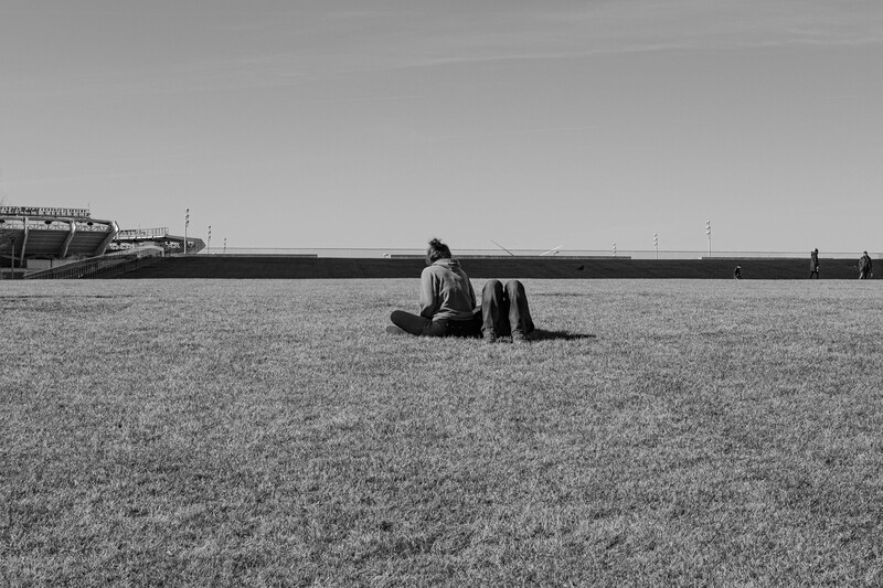 Person sitting alone in an expansive grain field under overcast sky, shot in black and white.