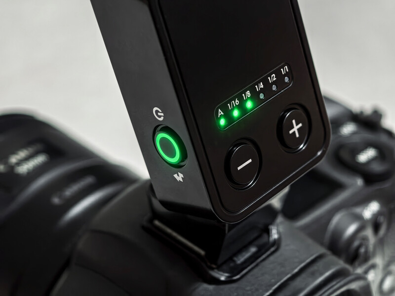 Close-up of a black wireless flash trigger mounted on a camera, with illuminated green LED indicators and control buttons visible.