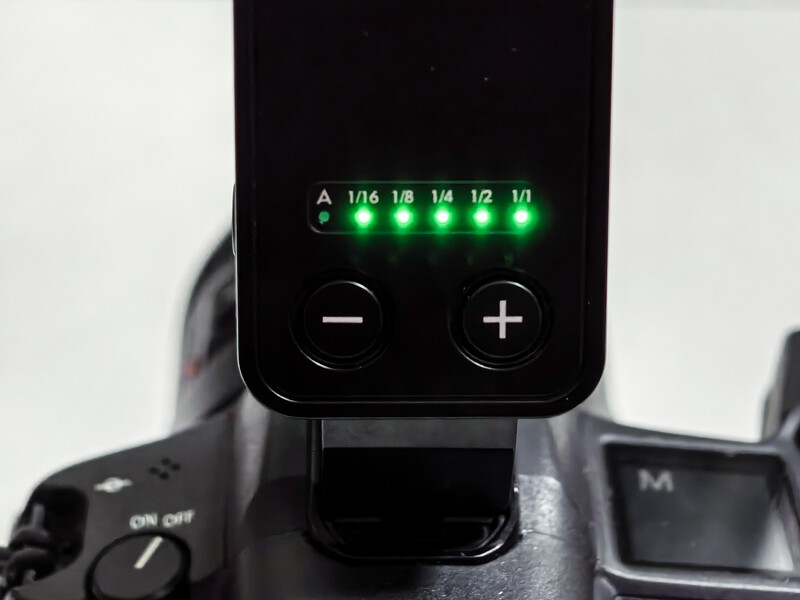 Camera flash unit displaying green LED aperture scale showing f-stops from f/16 to f/1.1, with minus and plus adjustment buttons below.