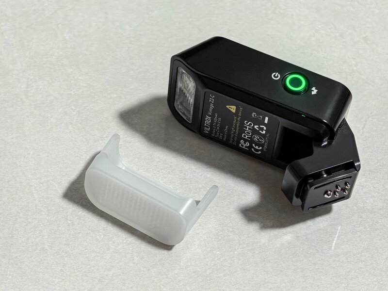 Black camera flash unit with white diffuser dome placed separately on light surface.