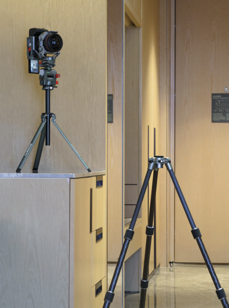Two professional video cameras mounted on tripods in an interior hallway with warm lighting.