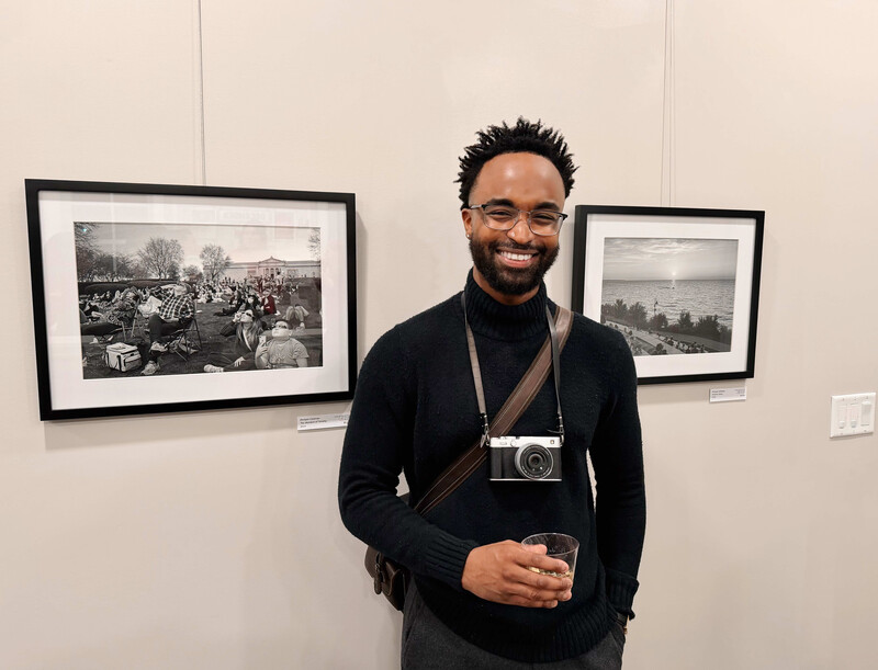 Photographer holding a vintage rangefinder camera, smiling in front of framed black and white photographs on a gallery wall.