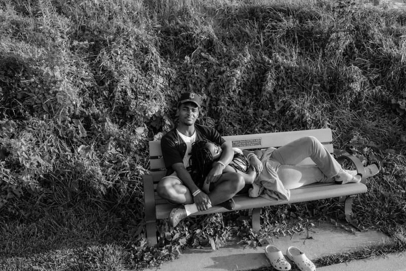 Man sitting on a bench surrounded by overgrown vegetation in black and white.