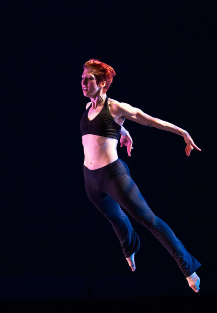 Athlete captured mid-jump against a black background, wearing a pink sports bra and blue leggings.