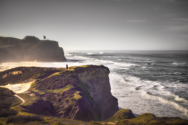 Solitary figure standing on moss-covered coastal cliffs overlooking turbulent ocean waves at golden hour.