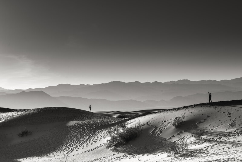 Two solitary figures stand on wind-sculpted sand dunes beneath layered mountains in monochrome.