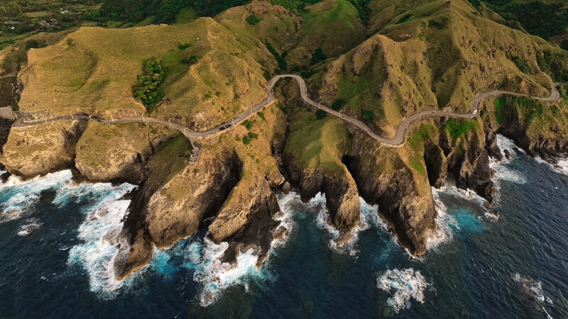 Aerial view of dramatic coastal cliffs with winding mountain road and crashing waves below.