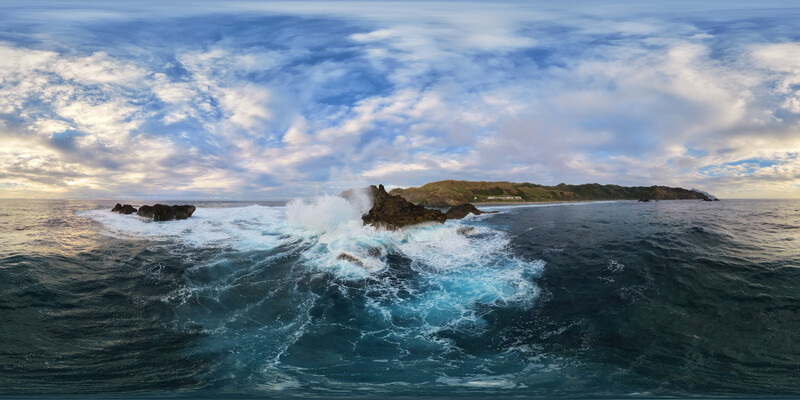 Rugged coastal landscape with large waves crashing against rocky islands beneath dramatic cloud-filled sky.