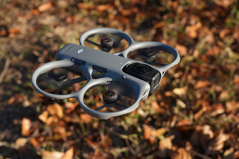 White and dark gray compact quadcopter drone hovering above autumn ground covered with fallen leaves.