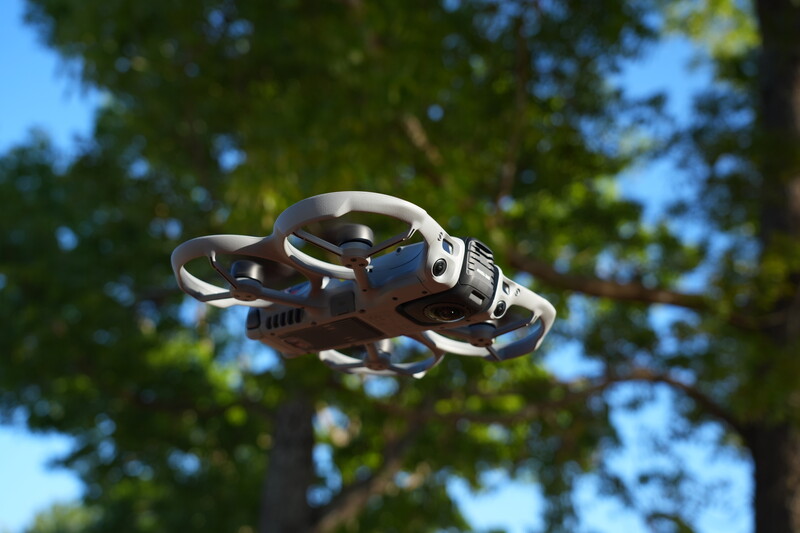 Compact quadcopter drone hovering against a bright blue sky with blurred green foliage in the background.