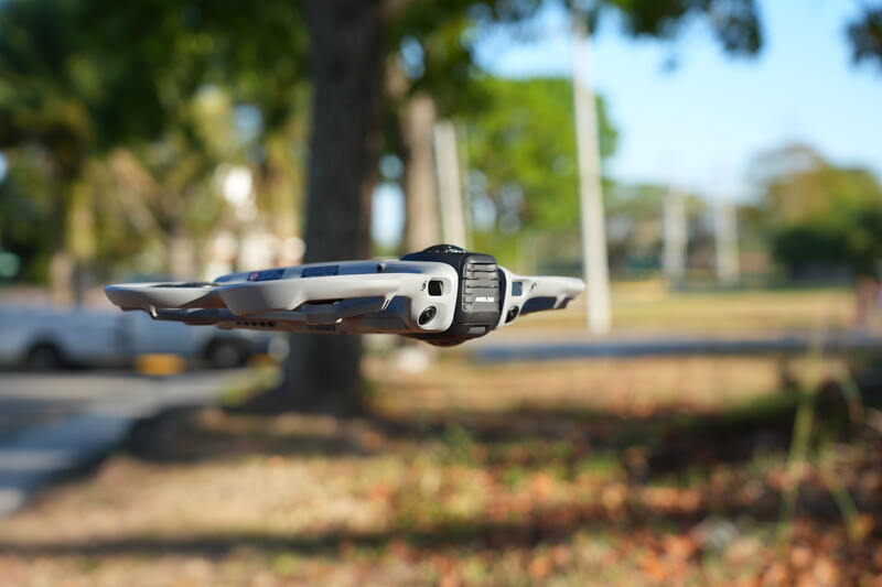 Drone hovering in sharp focus above a blurred park landscape with trees and pavement.