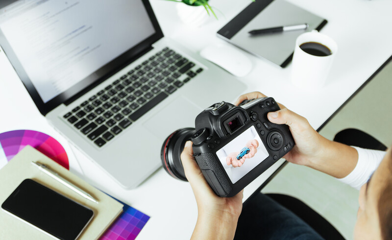 Photographer's hands holding a DSLR camera displaying an image on its rear LCD screen at a desk workspace.