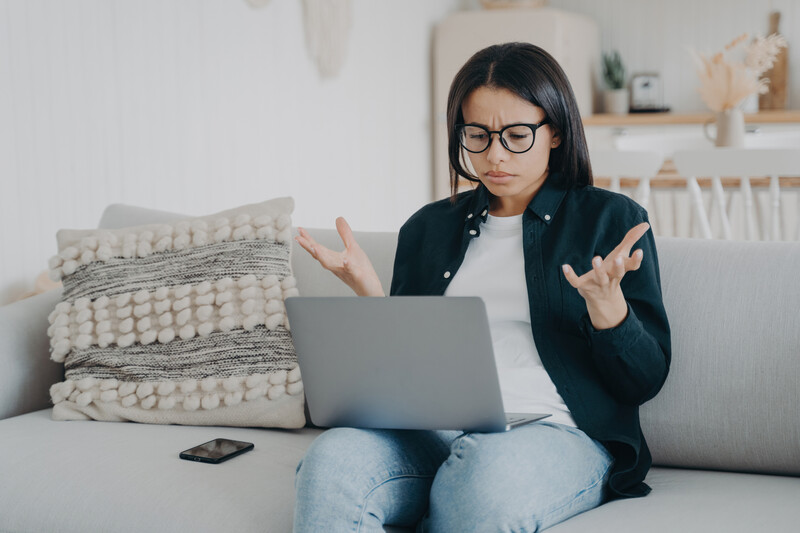 Woman in glasses and black blazer gestures expressively while viewing laptop on couch.