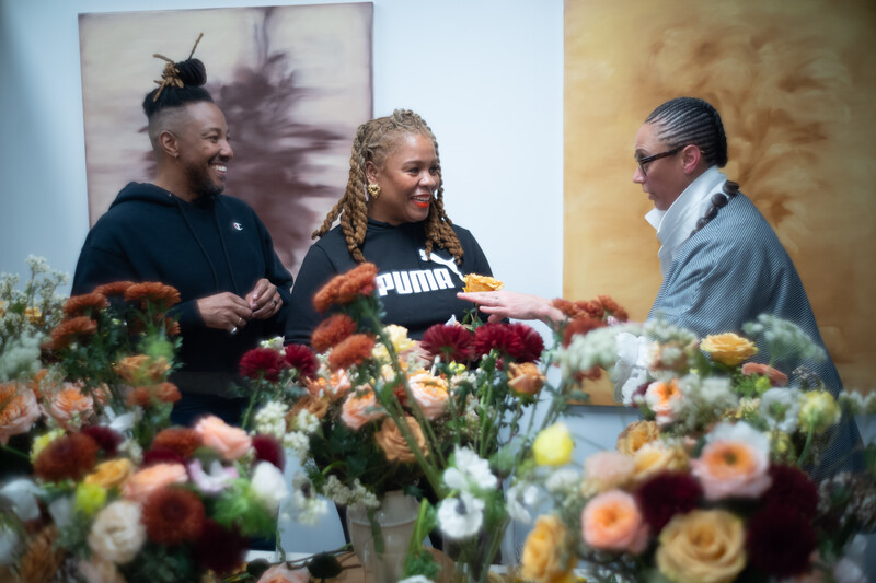 Three people conversing in front of colorful floral arrangements and artwork on gallery walls.