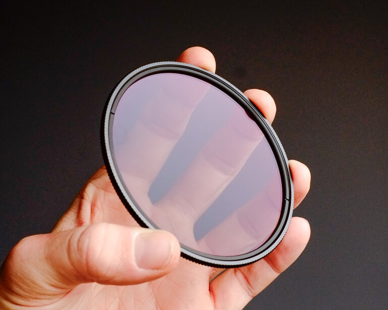 Hand holding a circular optical filter with a dark metal rim against a dark background.