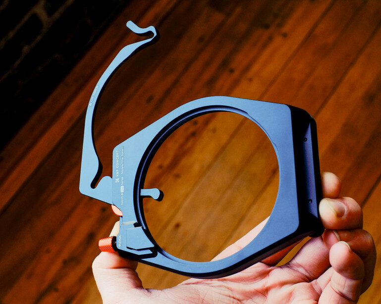Blue square lens filter holder with metal frame and adjustment arms, held in a hand over wooden surface.