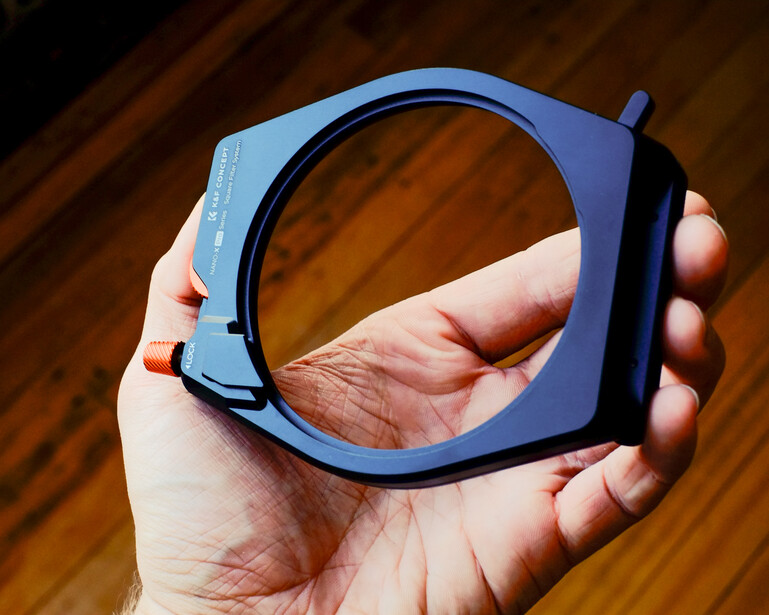Hand holding a blue square lens filter holder against a wooden surface.