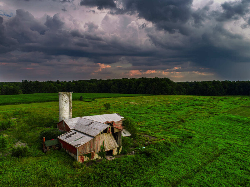 Abandoned wooden structure with metal chimney in vast green field under dramatic storm clouds.
