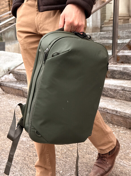 Person holding olive green camera backpack with shoulder strap while standing on outdoor steps.