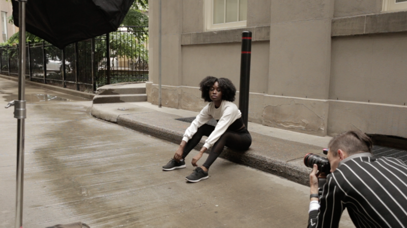 Photographer crouching while shooting a model sitting against an urban building facade.