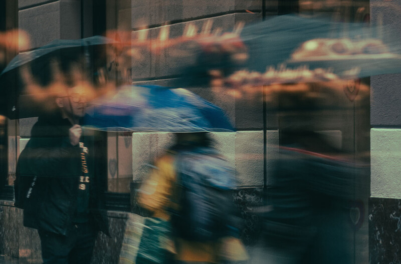 Multiple people in motion blur walking past glass storefronts and urban architecture.