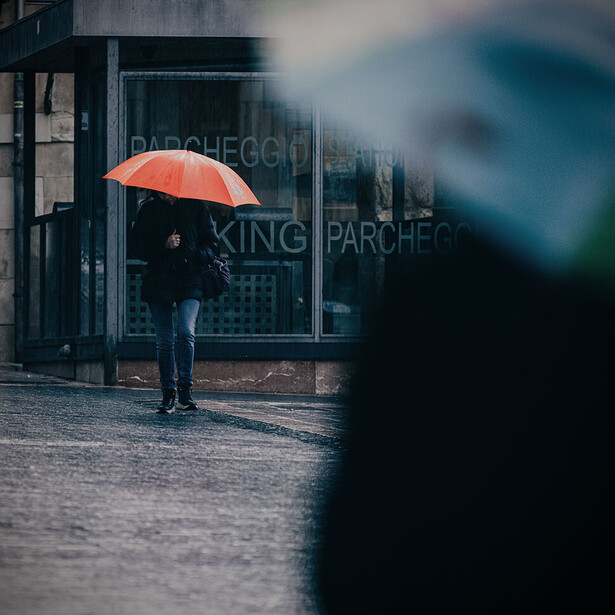 Person with orange umbrella standing on wet pavement outside a storefront during rain.