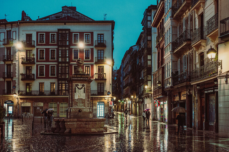Wet cobblestone street lined with illuminated historic buildings at dusk in a European city square.
