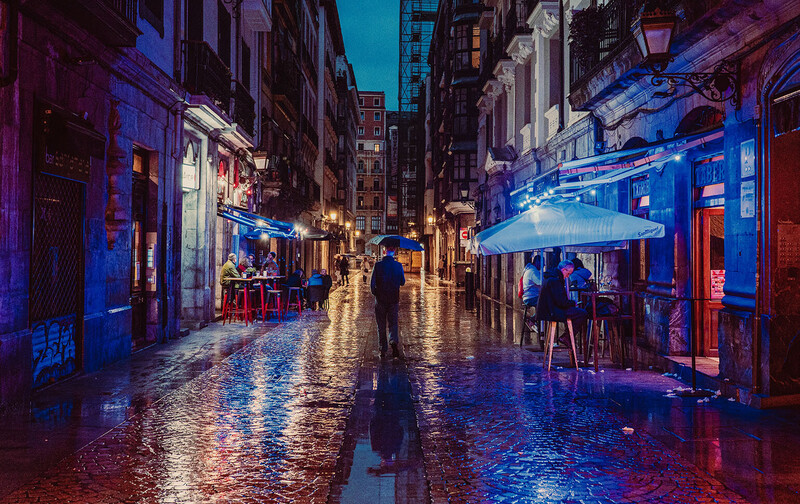 Wet cobblestone street at night illuminated by blue and purple neon lights reflecting off the pavement.