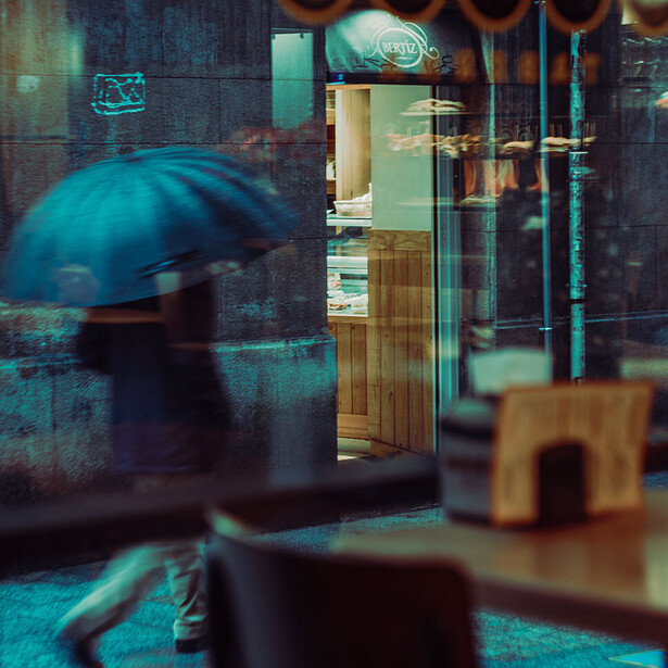 Person with umbrella walking past storefront with teal and cyan color cast overlay.