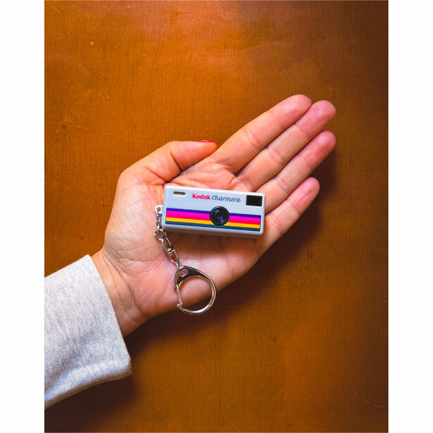 Hand holding a small colorful striped camera keychain against a warm brown background.