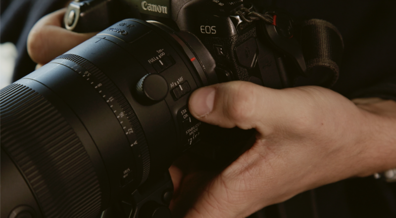 Photographer's hands holding a Canon EOS camera with a telephoto lens attached.