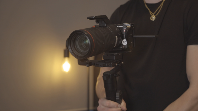 Filmmaker holding a DSLR camera with telephoto lens mounted on a gimbal stabilizer during indoor video production.