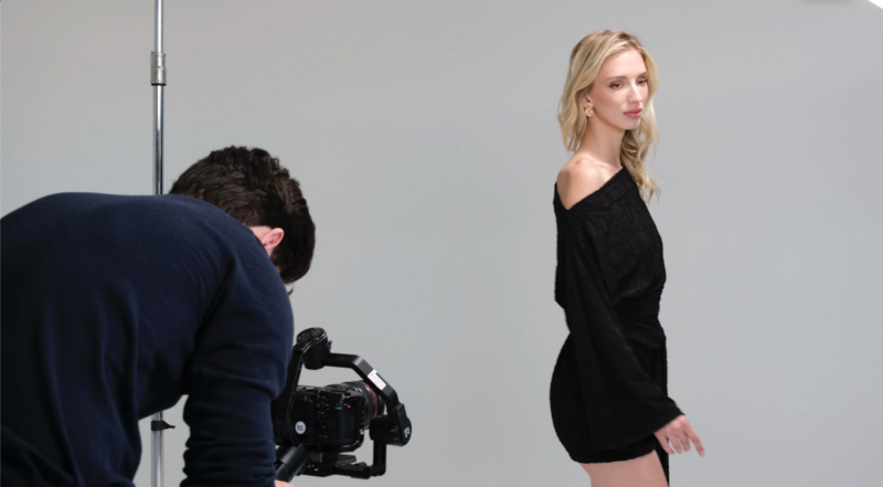 Cameraman filming a woman in black clothing against a gray studio backdrop with professional lighting.