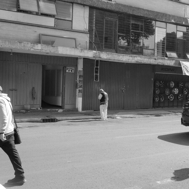 Black and white street scene with figures on an urban sidewalk in front of shuttered storefronts.