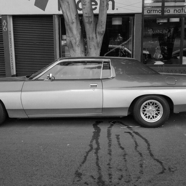 Black and white photograph of a vintage car parked on a wet street with storefronts in the background.