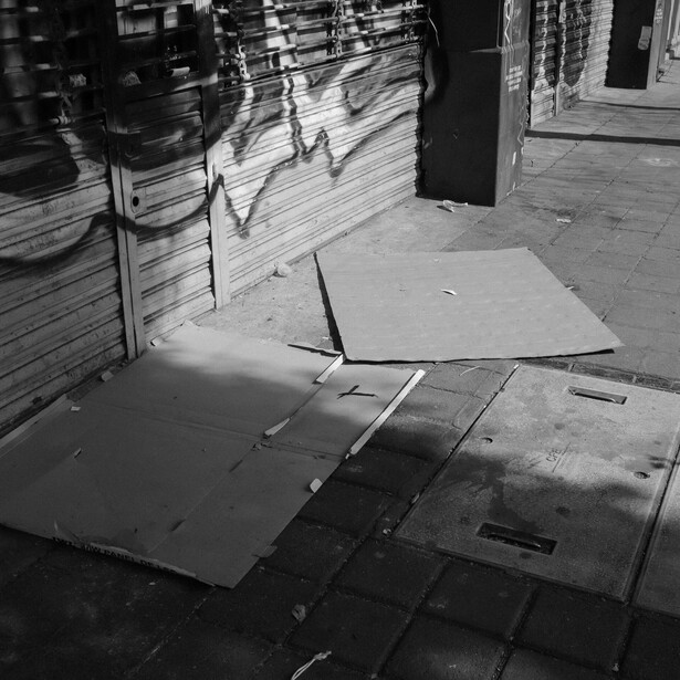 Black and white photograph of cardboard boxes arranged on a tiled street with dramatic shadow patterns from storefront elements.