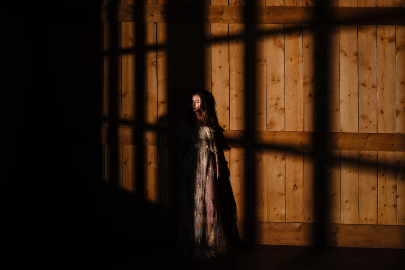 Woman in long dress standing against wooden wall with window grid shadow cast across her.