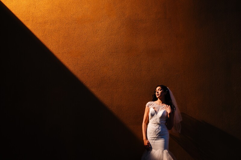 Woman in white mermaid gown standing in dramatic diagonal shaft of warm light against dark background.