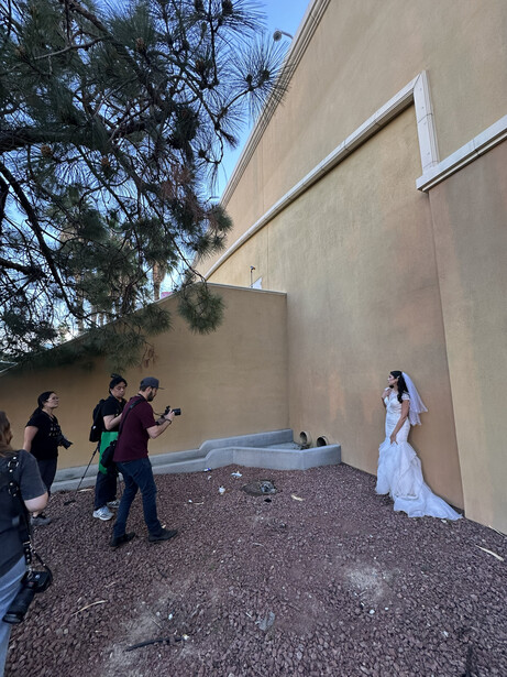 Bride in white dress posing for photos near concrete wall with crew and equipment.