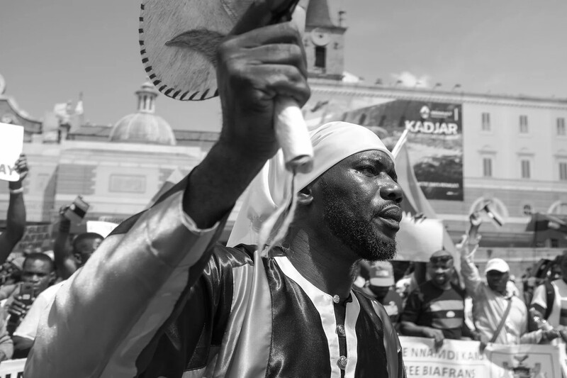 Man at outdoor rally raising fist with passion, wearing white cap and patterned shirt in black and white photograph.