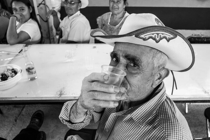 Older man in cowboy hat drinking from a glass at an indoor event, with spectators seated behind him.