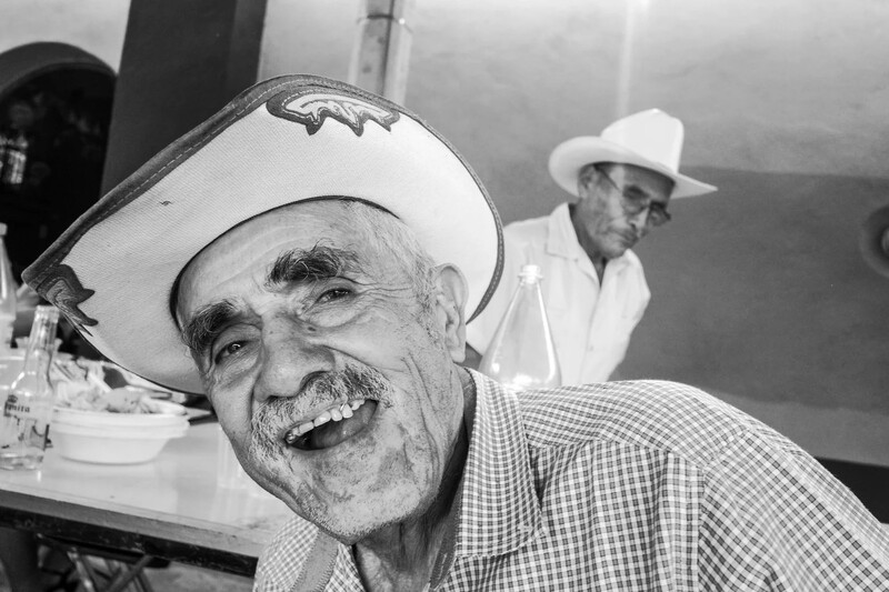 Older man in checkered shirt and cowboy hat smiling at camera, with another man in white shirt visible behind him.
