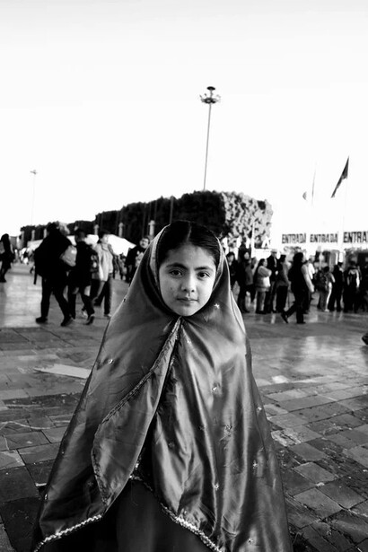 Young woman wearing a light-colored headscarf in a crowded public square, looking directly at camera.