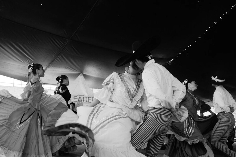 Black and white photograph of performers in period costumes dancing under a tent at night.