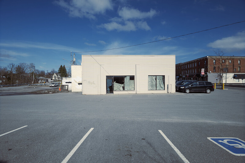 Modern white commercial building with large storefront windows in an empty parking lot under clear blue sky.