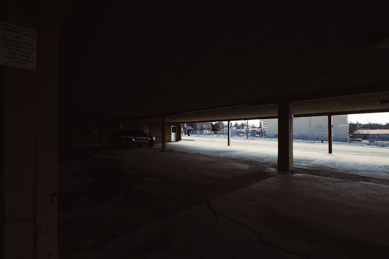 Dark interior parking structure with bright daylight streaming through large open windows revealing parked vehicles outside.