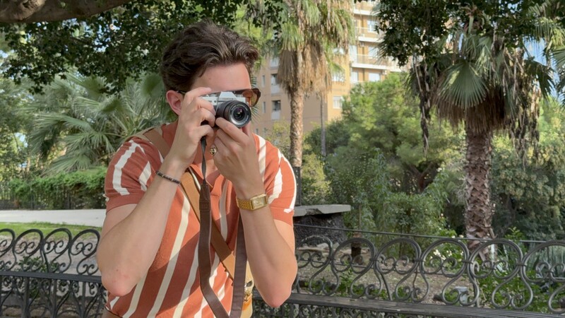 Photographer holding vintage rangefinder camera up to face while standing on a balcony overlooking palm trees and residential buildings.